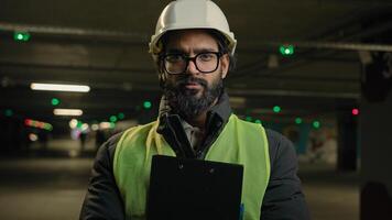 Portrait Indian man worker contractor mechanic guy in helmet and glasses posing with clipboard folder indoors parking factory warehouse manufacture. Arabian male builder engineer fitter in hard hat photo