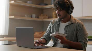 Indian man in headset look app pc browsing typing laptop write ideas questions notes review research sit at home kitchen. Arabian guy learn language improve knowledge self-education distant e-learning photo