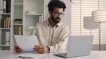 Focused man Arabian Indian ethnic businessman male employee manager writing notes business tasks data paperwork signing documents write on papers counting budget working with laptop computer in office photo