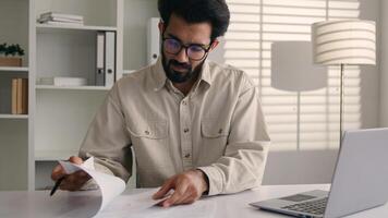 Focused man Arabian Indian ethnic businessman male employee manager writing notes business tasks data paperwork signing documents write on papers counting budget working with laptop computer in office photo