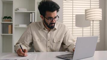 Focused man Arabian Indian ethnic businessman male employee manager writing notes business tasks data paperwork signing documents write on papers counting budget working with laptop computer in office photo