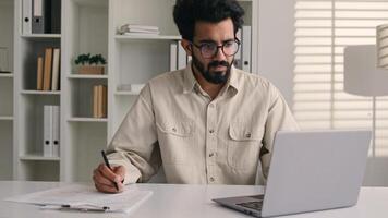 Focused man Arabian Indian ethnic businessman male employee manager writing notes business tasks data paperwork signing documents write on papers counting budget working with laptop computer in office photo