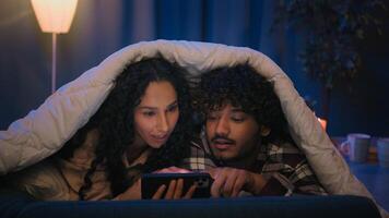 Gadget addict multiracial couple diverse man and woman scrolling mobile phone at night under blanket confused talking disagreement puzzled with smartphone error mistake shock bad news insomnia problem photo