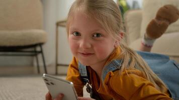 Close-up cute caucasian little kid girl lying on floor in living room using mobile phone application happy child playing telephone play online game on internet posing with smartphone at home photo