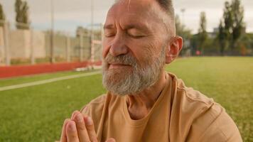 Old elderly Caucasian man stadium outside meditating on lawn sitting closed eyes smiling harmony retirement concentration wellness dreaming peaceful exercise balance fitness gymnastics posture sport photo