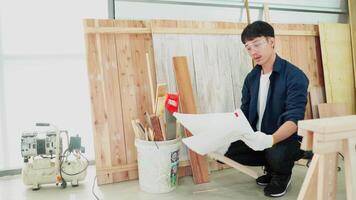 A man is kneeling down in front of a pile of wood and a bucket. He is holding a piece of paper and a pair of gloves. The scene suggests that he is working on a woodworking project video