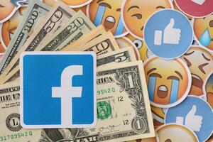 KYIV, UKRAINE - APRIL 20, 2024 Facebook logo and many usd money bills. Symbol of famous social network platform and dollar currency banknotes on lot of emoji reactions photo