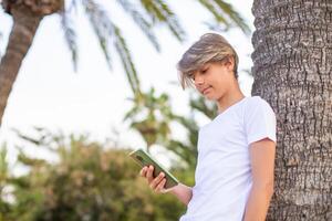 Close up handsome smiling teenager boy in a white t-shirt using mobile phone outdoors stands in a park near the big tree lunch time at summer.Side view from down angle,copy space. photo