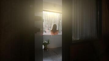Woman enjoying bath with forest view. A woman relaxing in a bathtub with a view of the forest through a large window, bathed in warm sunlight. video