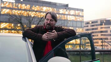 A man with a beard leans on his car, holding a smartphone, with illuminated modern buildings in the background at dusk. video