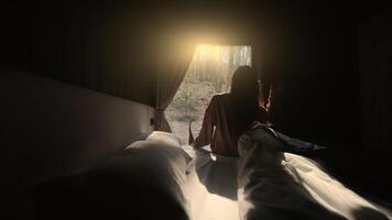A person sits on a bed in a dark room looking out of a window as the morning sun rises over a forest. The scene captures the calm and serene atmosphere of waking up in nature. video