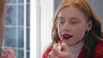A young woman with red hair is applying red lipstick while looking into a mirror. The image captures a moment of self-care and beauty, highlighting personal grooming and makeup application. video