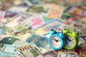 Small alarm clock on many banknotes of different currency. Background of time and money photo