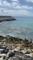Rocky coastline with breakwaters protecting the beach from waves. The blue sky and white clouds create a peaceful atmosphere. A bit of rubbish floating on the edge of the rocks video