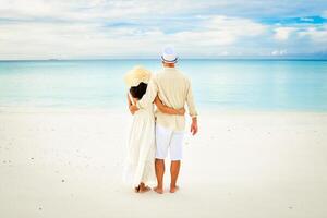 Back view beautiful tourist caucasian couple in white summer clothes and hats stands on a tropical beach and gazes at the turquoise ocean during their vacation time honeymoon photo