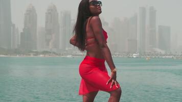 A young swarthy woman in a bikini and a light red dress walks barefoot on the rocks against the backdrop of the sea and skyscrapers of Dubai. video