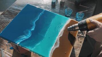 Artist uses dryer to create the effect of waves on the surface of the ocean in her epoxy resin painting. Close up. Slow motion video