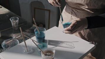 Women's hands are mixing blue paint. The artist is creating a painting in her studio using an unusual technique. Close up. Slow motion video