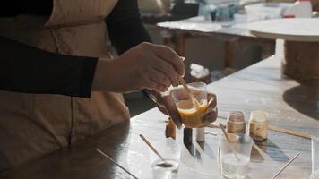 Woman is mixing honey-colored epoxy paint with a wooden stick in a plastic cup, in a creative studio, during a drawing masterclass. Close up. Slow motion video