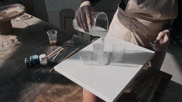 Craftswoman is pouring viscous, transparent resin from a large container into smaller ones. She is creating a base for the paint. Mid shot. video