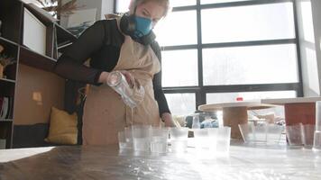 An artist, wearing a respirator, experiments with epoxy resin in her studio. She pours the resin into several transparent plastic cups. Mid shot. Slow motion video