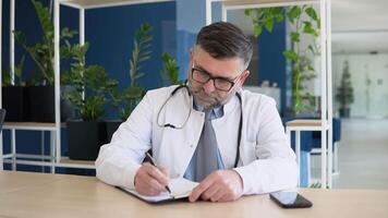 Focused middle aged senior head doctor in white medical coat and glasses sitting at workplace, writing notes in paper journal video
