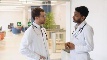 Medical multiethnic staff having discussion in a hospital hallway. Indian and caucasian smiling doctors in white coat working in a medical clinic. Two hospital workers discussing on laboratory test video