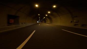 First person view, car is driving through tunnel in mountains. Light at end of tunnel becomes visible as car moves forward. Interior lights illuminate tunnel walls, guiding the way video