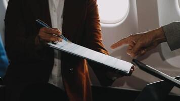 Two businessman passengers sitting in airplane cabin and working on laptop together. Businessman in brown suit showing project on notebook to businessman with eyeglasses. Business and travel concept. video
