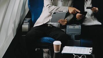 Two businessman passengers sitting in airplane cabin and working on laptop together. Businessman in brown suit showing project on notebook to businessman with eyeglasses. Business and travel concept. video