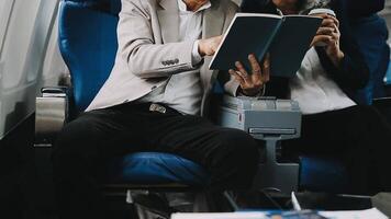 Two businessman passengers sitting in airplane cabin and working on laptop together. Businessman in brown suit showing project on notebook to businessman with eyeglasses. Business and travel concept. video
