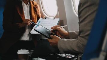 Two businessman passengers sitting in airplane cabin and working on laptop together. Businessman in brown suit showing project on notebook to businessman with eyeglasses. Business and travel concept. video