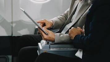 Two businessman passengers sitting in airplane cabin and working on laptop together. Businessman in brown suit showing project on notebook to businessman with eyeglasses. Business and travel concept. video
