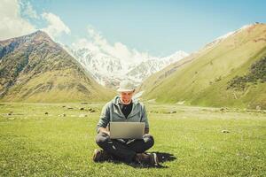 Young caucasian male person digital nomad sits in mountains meadow filed with laptop types with smile in his face. Remote work and blogging while traveling concept photo