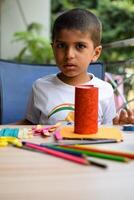 Smart Indian little boy performing best out of waste activity with different colourful objects during the summer vacations, Cute Indian Kid doing best our of waste activity at home on wooden table photo