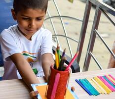 Smart Indian little boy performing best out of waste activity with different colourful objects during the summer vacations, Cute Indian Kid doing best our of waste activity at home on wooden table photo