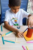 Smart Indian little boy performing best out of waste activity with different colourful objects during the summer vacations, Cute Indian Kid doing best our of waste activity at home on wooden table photo