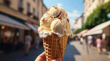 A hand holding a creamy ice cream cone on a sunny day. The vanilla gelato in a waffle cone is topped with nuts, against a vibrant city street backdrop with people, capturing the essence of summer photo
