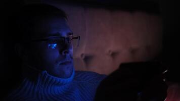 Lonely man in glasses sitting on bed in bedroom and using phone at night. Face illuminated from smartphone display. Surfing social network before sleep photo