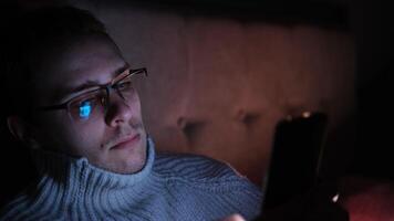 Lonely man in glasses sitting on bed in bedroom and using phone at night. Face illuminated from smartphone display. Surfing social network before sleep photo