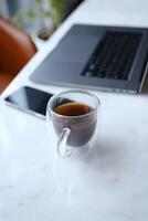 workplace with phone laptop and transparent cup of coffee in home office on white marble desk with brown leathern chair and green tree at background photo