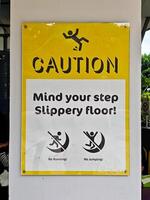 Bright yellow caution sign with stick figure and text Mind your step Slippery floor posted on a swimming pool area emphasizing safety precautions and warning against running or jumping. photo