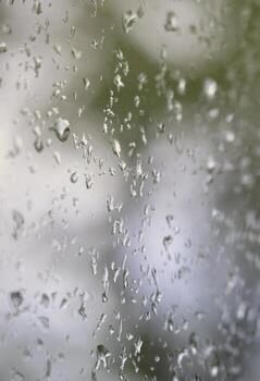 A photo of rain drops on the window glass with a blurred view of the blossoming green trees. Abstract image showing cloudy and rainy weather conditions