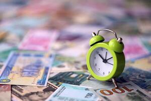 Small alarm clock on many banknotes of different currency. Background of time and money photo