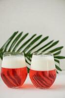Cocktail in transparent glasses. Layers of jelly and whipped cream. Strawberry cocktail on a white background with a leaf of palm tree.Exotic. photo