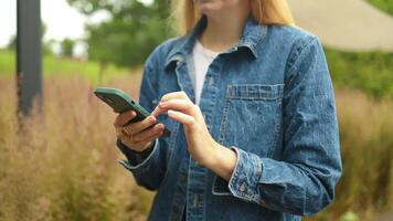 Happy pretty girl student hands user holding cell phone using education tech apps looking at smartphone texting messages standing outside. Mobile online applications concept. video