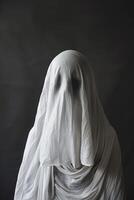 Ghostly figure draped in white, standing against dark backdrop. The minimalist composition and mysterious ambiance make it perfect for Halloween themes and horror projects. Copy space. photo