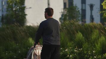 Rear view of a father with a stroller walking along greenery at the courtyard. Stock footage. Man and his baby child walking outdoors on a summer day. video