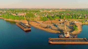 Bird's-eye view. Scene.The river on which there are ships for construction with cargo on them and a bright city with green summer trees against a blue sky is visible. video