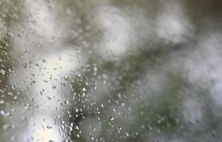 A photo of rain drops on the window glass with a blurred view of the blossoming green trees. Abstract image showing cloudy and rainy weather conditions